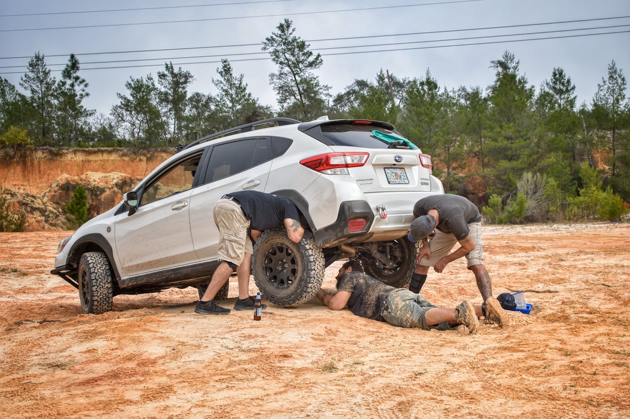 gallery-05 Three men work together to repair a vehicle in an outdoor setting, surrounded by trees and rocky terrain.