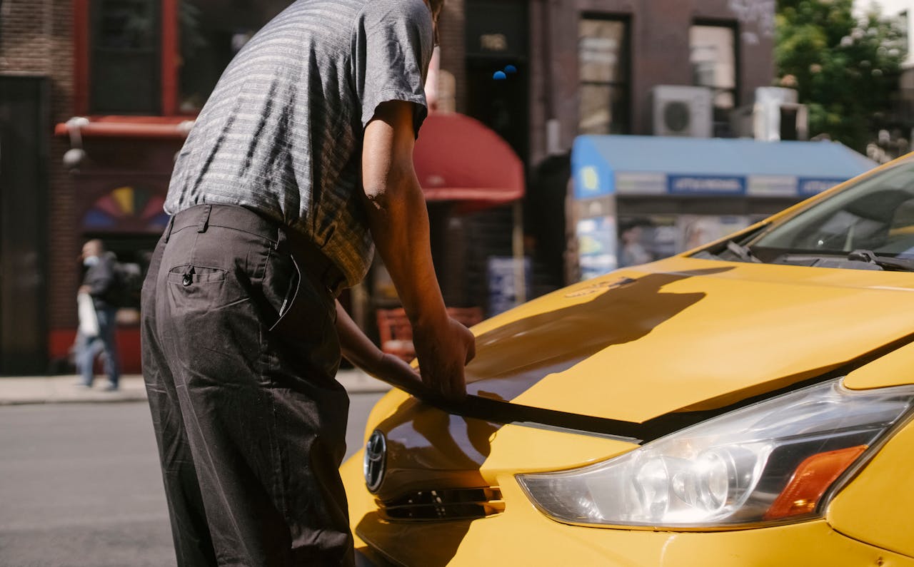 gallery-06 Man inspecting open hood of a yellow taxi on city street.